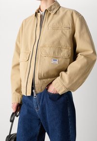 Beżowa kurtka zapinana na zamek z dwoma przednimi kieszeniami i małą kieszonką na piersi, o gładkiej fakturze i skróconym fasonie. Nosi się ją na niebieskie dżinsy.