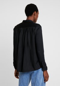 Schwarze Bluse mit Falten-Design am Rücken, Kragen und Knopfmanschetten, kombiniert mit hellblauen Jeans. Glatte Textur und minimalistischer Stil.