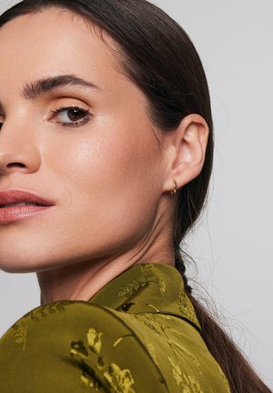 Złota kolczyk w kształcie kółka, gładkie wykończenie. Modelka nosi zielony top w wzory z kołnierzykiem, z kwiatowymi detalami. Karnacja skóry neutralna, delikatny makijaż.
