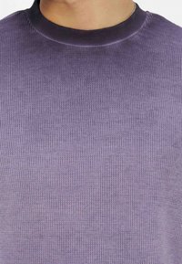 Κοντινό πλάνο ενός ατόμου που φοράει μοβ πλεκτό τοπ με μαύρο στρογγυλό γιακά και ασημένια αλυσίδα για λευκό.