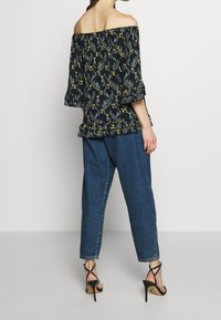 Blusa floral negra con patrones amarillos y verdes, de tirantes caídos, con dobladillo con volantes, combinada con unos vaqueros azul oscuro de cintura alta y tacones negros.