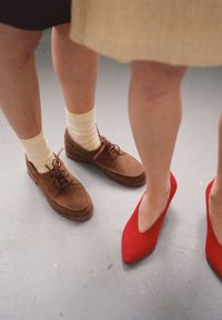 Scarpe in suede marrone con lacci, abbinate a calzini a scacchi gialli. Tacchi a punta rossa accanto a loro, su un pavimento grigio.