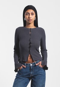 Gray ribbed long-sleeve top with scalloped edges, front buttons, and a cropped cut, paired with high-waisted blue jeans.
