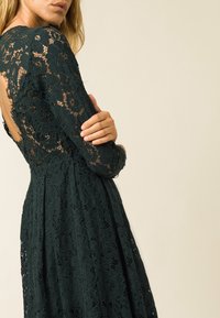 Donkergroene kanten jurk met lange mouwen en een diepe opening op de rug. Bloemenpatroon door de jurk en een uitlopende rok met een textuurafwerking.