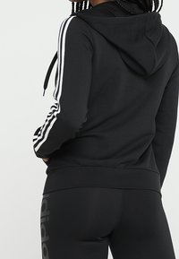 Sudadera negra con rayas laterales blancas, capucha y puños acanalados. Combinada con leggings negros que tienen un logo en el lateral. Textura de tela suave.