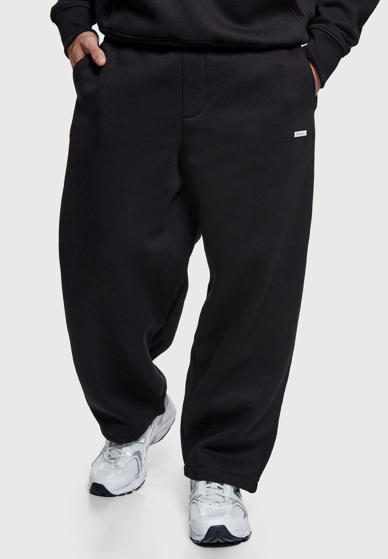 Pantalon de survêtement noir oversize en tissu doux, doté d'une coupe large, d'une taille élastique et de poches latérales, porté avec des baskets blanches.