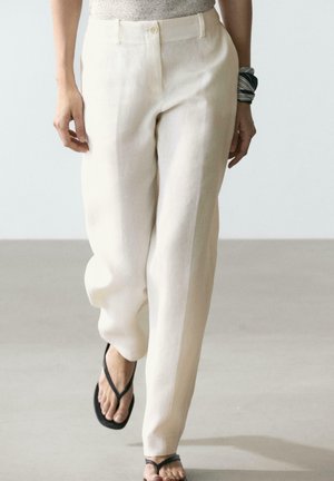 Donna che cammina indossando pantaloni a vita alta color bianco sporco, sandali infradito neri e un foulard bianco e nero al polso destro.