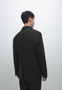 Dunkelgrauer Wollblazer mit klassischem Revers, strukturierten Schultern und tailliertem Schnitt. Besitzt einen Schlitz auf der Rückseite und knöpfbare Ärmelabschlüsse.
