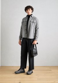 Grå ullskjortejakke, svart genser med høy hals, svarte bukser, grå joggesko og en liten svart crossover-veske i et minimalistisk miljø.