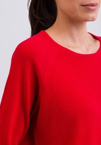 Pullover rosso in maglia con scollo rotondo e maniche lunghe a raglan. Il tessuto sembra morbido, con una texture liscia e una leggera lucentezza.