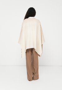 Kremfarget poncho med myke horisontale striper i lys brun, med drapert design og åpen front. Lett, teksturert stoff med avslappet passform.