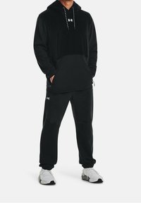 Uomo che indossa un cappuccio nero Under Armour e pantaloni jogger neri abbinati con scarpe da ginnastica bianche, in piedi con le mani in tasca.