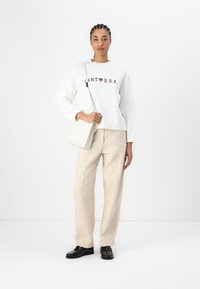 Sweatshirt blanc avec le texte « CANT U.S.A. » en navy, pantalon beige ajusté, chaussures noires et un petit sac blanc. Tissu lisse, coupe décontractée.