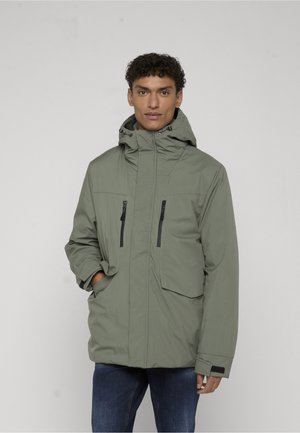 Homme portant une veste à capuche vert olive avec des poches poitrine zippées et des poches latérales à rabat, debout devant un fond uni.