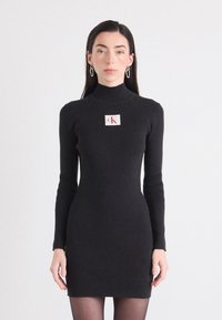 Model u uskom crnom haljini s rebrastim uzorkom, dugim rukavima i visokim ovratnikom, s CK logotipom na prsima. Jednobojna bijela pozadina.
