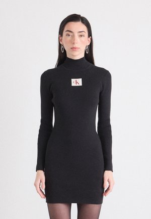 Modelo vistiendo un vestido negro ajustado de canalé con mangas largas y cuello alto, que presenta un logo de CK en el pecho. Fondo blanco liso.