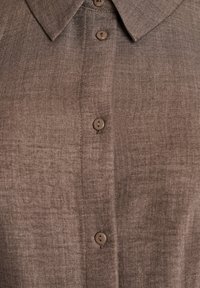 Hnědá košile na knoflíky z texturované tkaniny, s límečkem a šesti odpovídajícími knoflíky vpředu pro zapínání a detail.