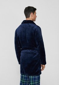 Tmavě modrý plyšový župan s šálovým límcem, páskem v pase a dlouhými rukávy. Jemná textura s hladkým povrchem.