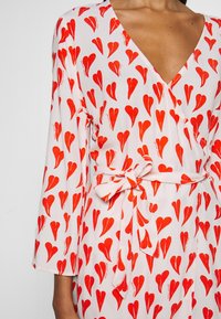 Frau trägt ein weißes Wickelkleid mit roten herzförmigen Mustern und einer gebundenen Schleife an der Taille, mit einem V-Ausschnitt und drei Viertelärmeln.