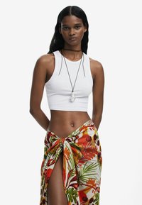 Top corto bianco abbinato a un sarong colorato e fantasioso con disegni tropicali in rosso, verde e arancione, legato in vita.