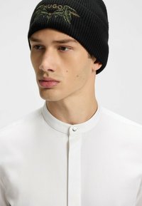 Black knitted beanie with green embroidered logo, paired with a white, collared shirt featuring a hidden button closure.