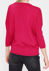 Fuchsia stickad tröja med avslappnad passform, trekvartslånga ärmar och ribbad struktur, i kombination med svarta jeans, visad bakifrån.