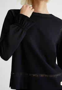 Sudadera negra de corte holgado, con puños elásticos, borde de encaje en el dobladillo y un acento de logotipo en el lateral. Textura suave.