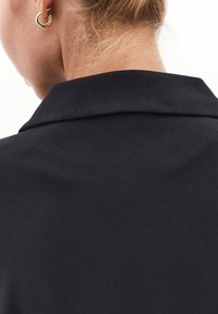 Černý top s límečkem vyrobený z hladké tkaniny, s čistým výstřihem a jemnou texturou, s viditelným zlatým kruhovým náušnicem.