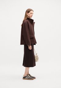 Tmavě hnědá oversized bunda s vysokým límcem, odpovídající sukně a šedé tenisky. Outfit doplňuje ramenní taška ve světlé barvě.