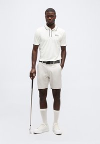Homme portant un polo blanc, un short de couleur claire, des chaussettes blanches et des baskets, tenant un club de golf et se tenant devant un fond blanc uni.