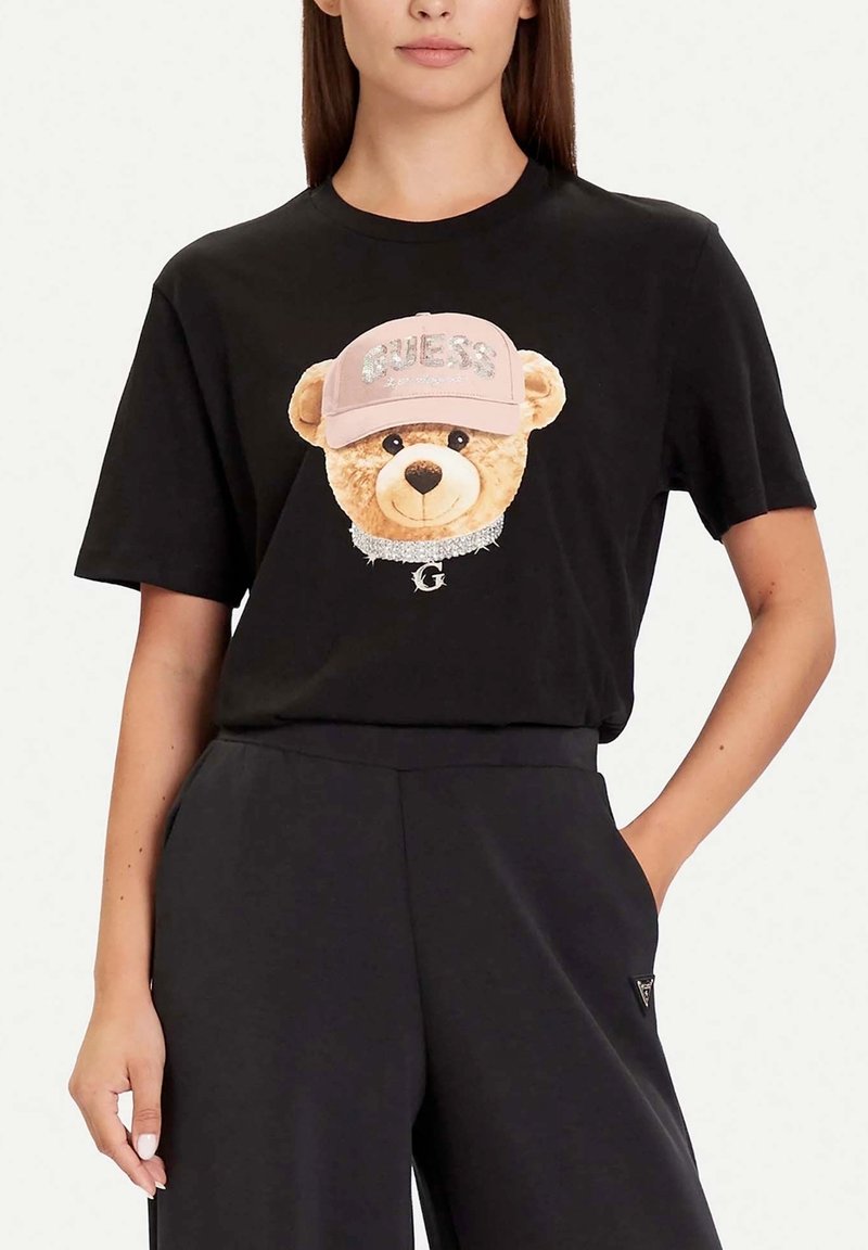 Camiseta negra de algodón con una imagen de un oso de peluche de felpa que lleva una gorra rosa con el estampado "GUESS" y un detalle de collar brillante.