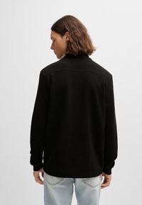 Pullover preto de mangas longas com acabamento texturizado, apresentando um colarinho alto e um corte reto, combinado com jeans de denim claro.