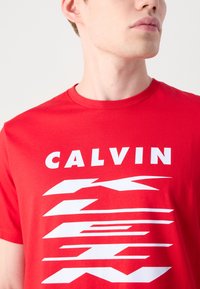 Raudona medvilninė marškinė su ryškiu baltu tekstu "CALVIN" ir dinamiškomis linijomis modernesniame dizaine, apvali apykaklė ir trumpomis rankovėmis.