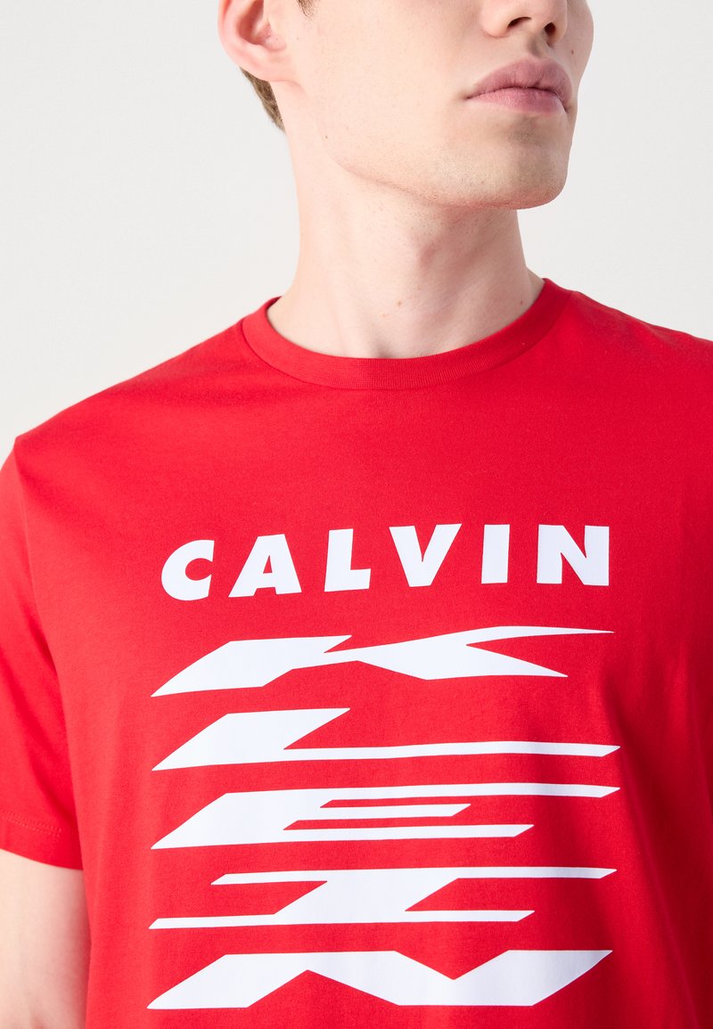 T-shirt en coton rouge avec le texte en gras "CALVIN" en blanc et des lignes dynamiques dans un design moderne, col rond et manches courtes.