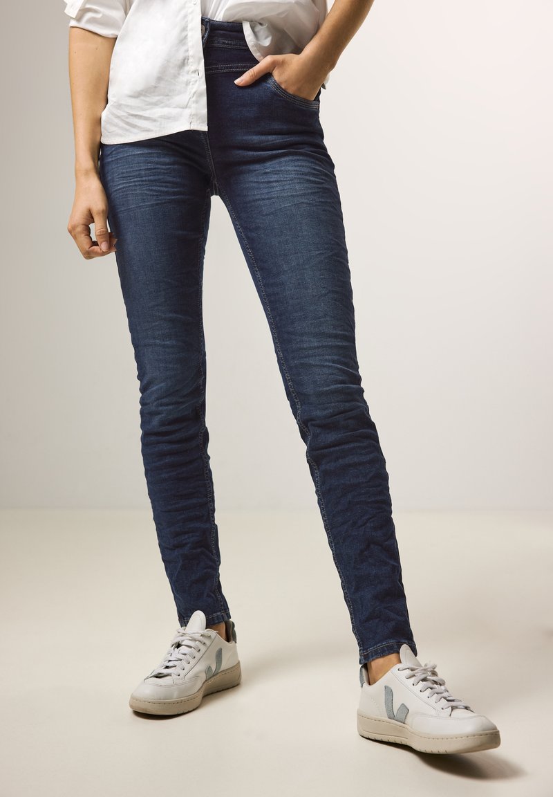 Jeans skinny bleu foncé en denim, taille haute avec poches avant, associés à des baskets blanches avec des accents gris et noir.