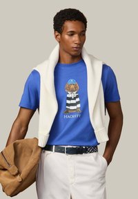 Mann trägt ein blaues T-Shirt mit Hundemotiv, einen weißen Pullover über den Schultern, weiße Hosen und hält eine beigefarbene Wildlederjacke.