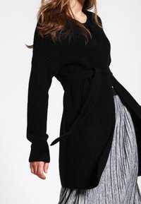 Žena má na sobě dlouhý černý pletený kardigan se páskem, který nosí přes šedou plisovanou sukni, na jednobarevném pozadí.