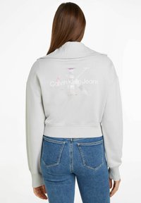 Grå beskuren sweatshirt med en bred krage, som har en stor iriserande "CK"-logotyp och texten "Calvin Klein Jeans" på baksidan.