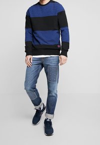 Sweter crewneck w czarno-niebieskim paskowym wzorze, w połączeniu z jasnoniebieskimi dżinsami z teksturowanymi łatami. Ciemne sneakersy dopełniają całość.