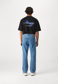 Svart T-shirt med grafisk text "L'HOMME DE LA NUIT" och blåa accenter, kombinerad med ljusblå raka jeans och vita sneakers.