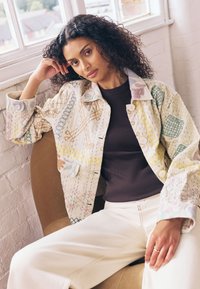 Mehrfarbige bestickte Jacke mit komplizierten Mustern auf hellem Stoff, kombiniert mit einem dunklen Oberteil und cremefarbenen Hosen, sitzend auf einem Stuhl.