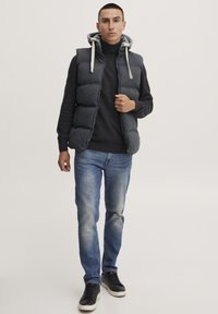 Svart puffervest med grå hette, brukt over en mørk genser og blå jeans, kombinert med svarte sneakers med hvit såle.