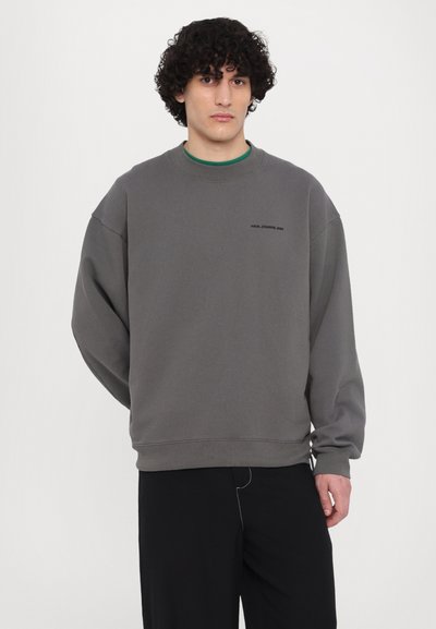 Grå sweatshirt med rund hals, nedfelte skuldre, ribbestrikkede mansjetter og en liten svart logo på brystet; kombinert med svarte bukser.