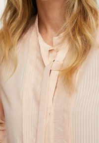 Pfirsichfarbene Bluse mit vertikalen Streifen, einem Bindekragen und strukturiertem Stoff. Der weiche Fall und der dezente Glanz verstärken das Design.