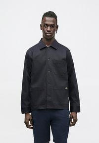 Chaqueta azul marino abotonada, hecha de tela con textura suave, con un bolsillo en el pecho, dos bolsillos frontales y un detalle de logo.