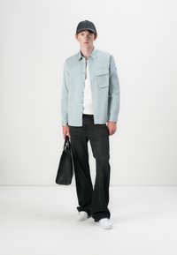 Camisa azul clara, suéter branco, jeans pretos e ténis brancos. Segurando uma mala preta. A camisa possui um bolso no peito e botões.