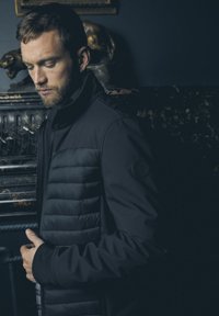 Zwarte pufferjack met een gladde stof aan de bovenkant en een gecapitonneerde textuur aan de onderkant; heeft een hoge kraag en een logo-patch op de mouw.