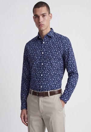 Jeune homme portant une chemise bleue à motifs floraux rentrée dans un pantalon beige avec une ceinture marron foncé, debout avec une main dans la poche.