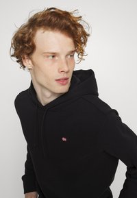 Jong persoon met krullend rood haar, gekleed in een zwarte hoodie met een klein rood-wit vlagembleem, kijkt naar de zijkant tegen een effen achtergrond.