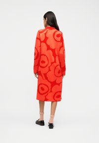 Γυναίκα που φοράει ένα μακρύ κόκκινο φόρεμα με μεγάλα πορτοκαλί λουλουδένια σχέδια, ροζ κάλτσες και μαύρα μοκασίνια, που στέκεται μπροστά σε ένα απλό φωτεινό φόντο.